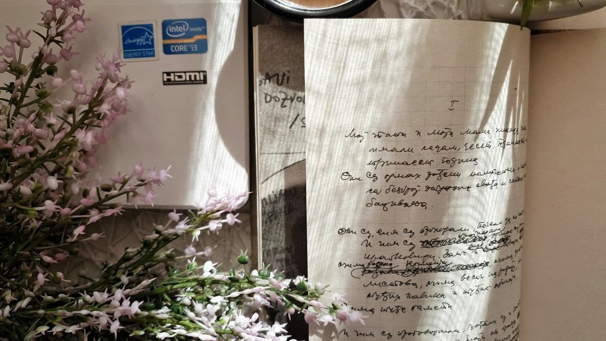 A creative workspace with notebook, coffee, laptop, and pink flowers.