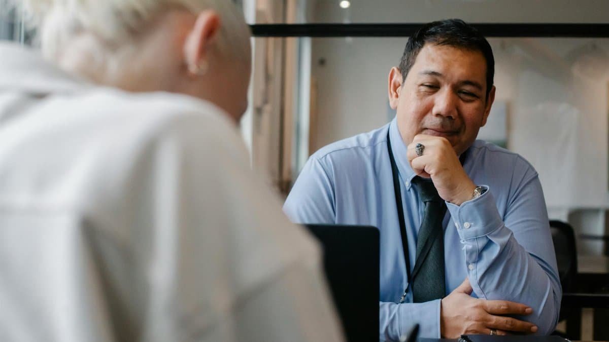 Confident ethnic male employee sitting at contemporary workplace and looking satisfied while young male client sitting in front