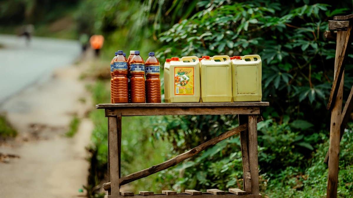 Rural roadside stall featuring bottles and jerrycans of oil and juice with a lush green backdrop.