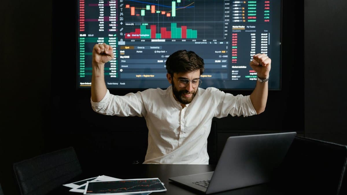 Businessman celebrates stock market success with hands raised in excitement at a trading desk.