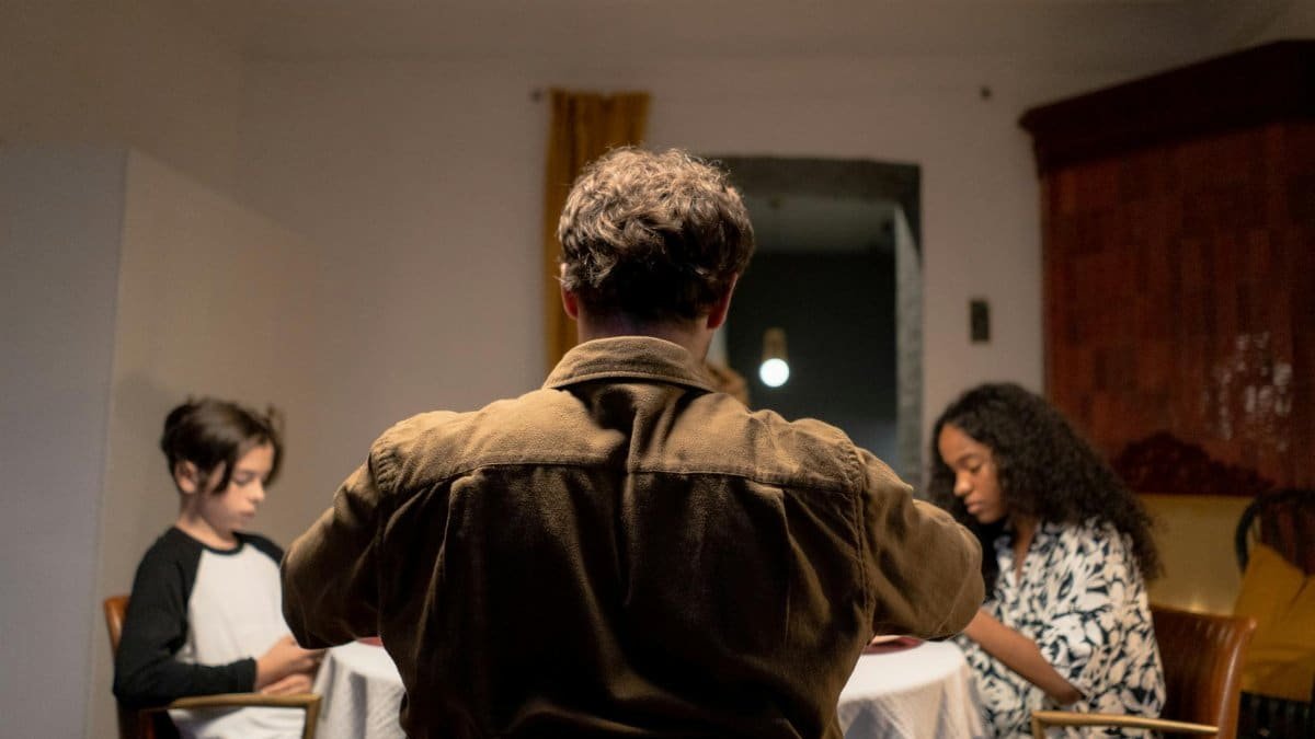 A family gathers around a dining table, featuring a father and two children in an intimate setting.