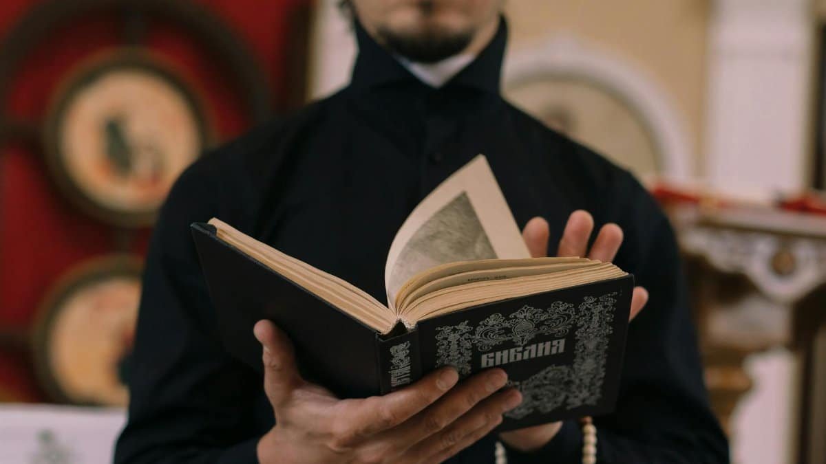 A religious figure in a black robe reading from a decorated sacred book indoors.