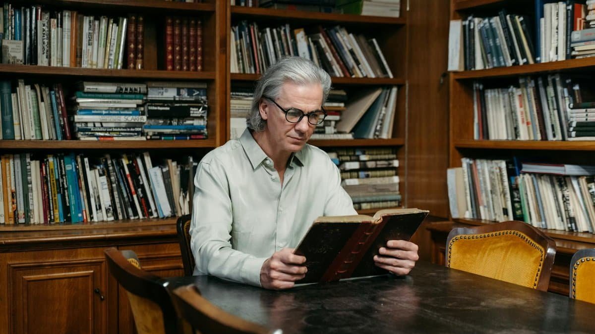 Elderly man reading in a library surrounded by bookshelves, promoting wisdom.