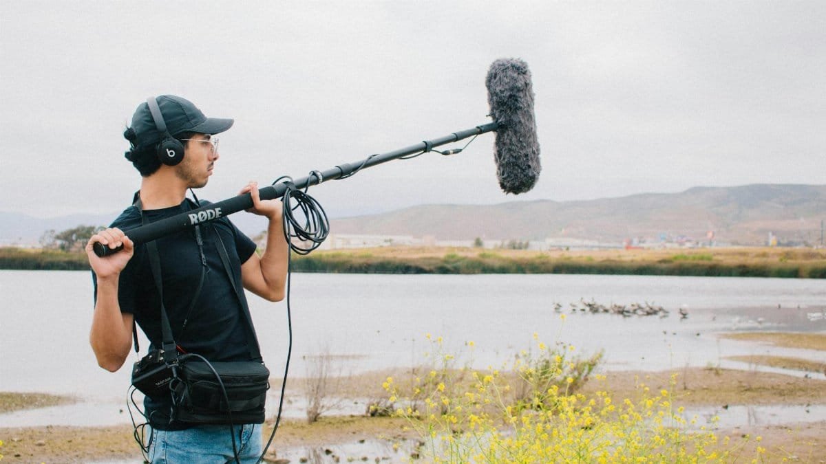 A sound technician operates a boom microphone near a lake with scenic hills in the background.
