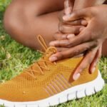 Close-up of athlete holding ankle with running shoe on grass, indicating pain or injury.