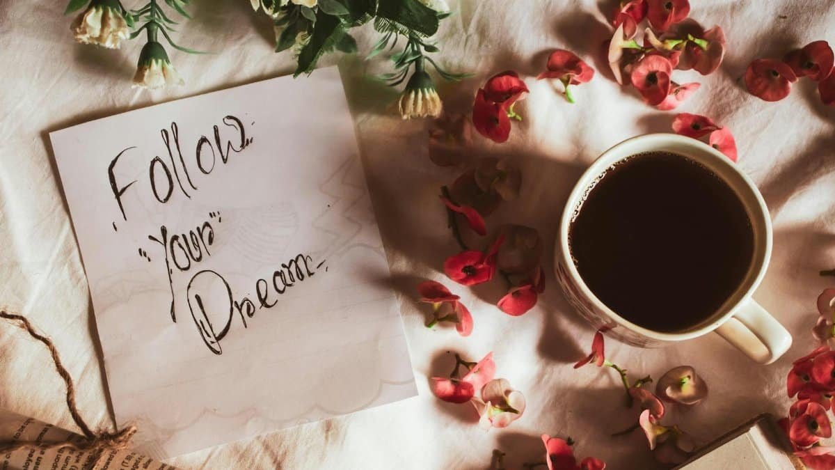 Top view of Follow Your Dreams inscription on paper placed on textile near mug of hot beverage and flower petals