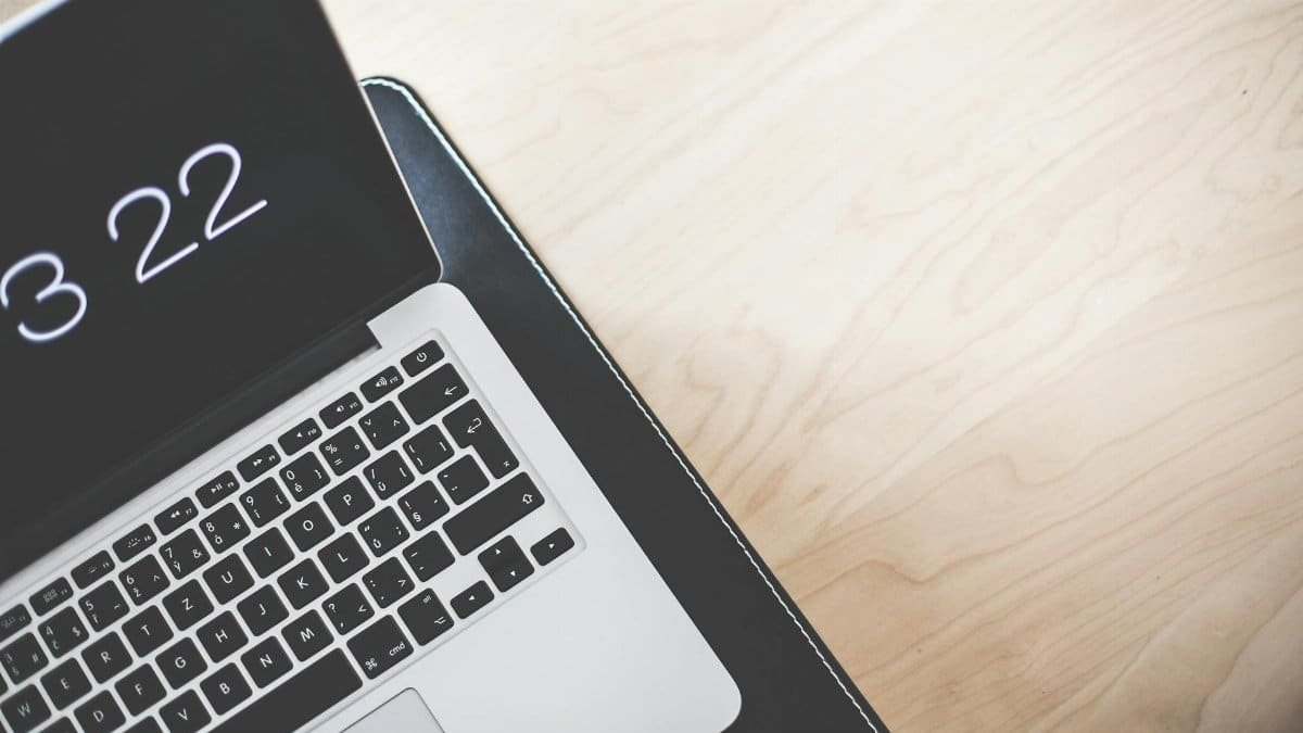 A minimalist workspace featuring a laptop with an open screen showing the time, on a light wooden desk.