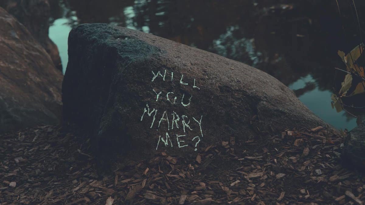 Chalk-written marriage proposal on a rock by a lake in a serene forest setting.