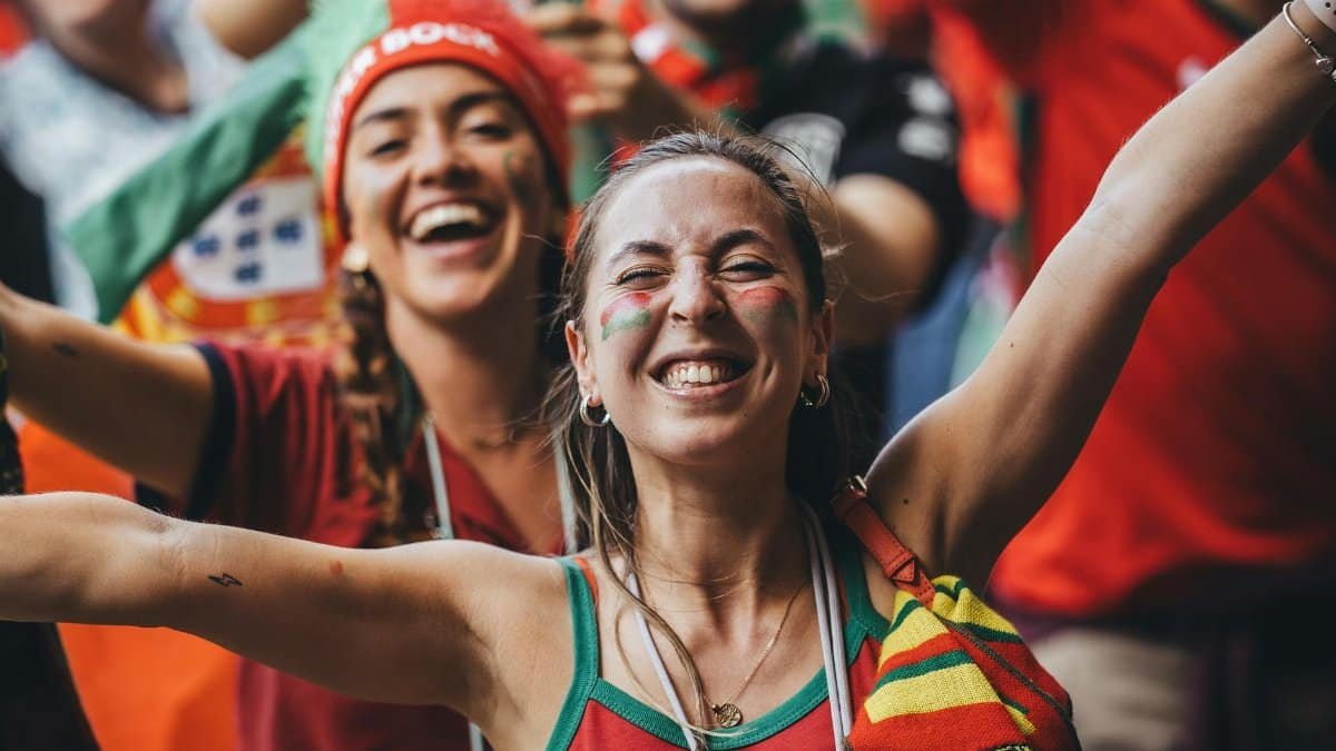 Excited women fans celebrate with flags and smiles during a sports event outdoors.