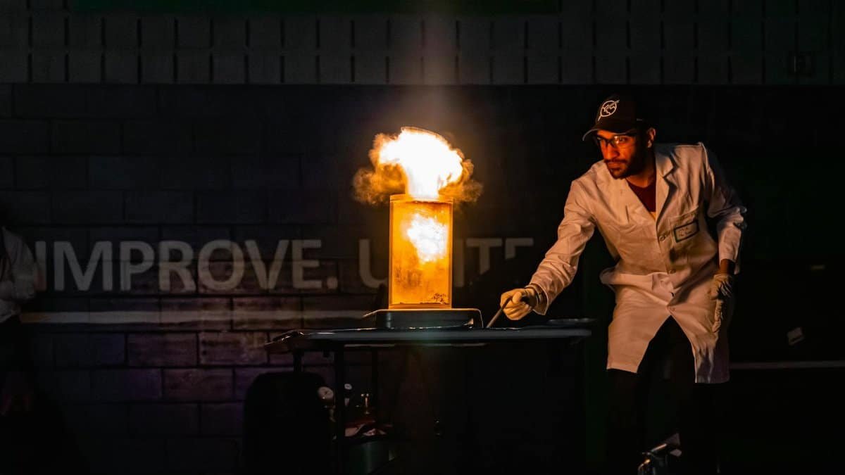 A scientist in a lab coat demonstrates a dramatic fire explosion experiment indoors.