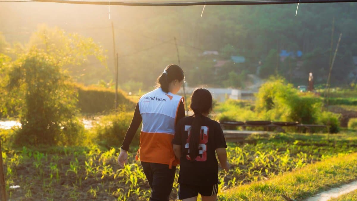 Volunteer in World Vision vest walking with child in scenic rural landscape at sunset.
