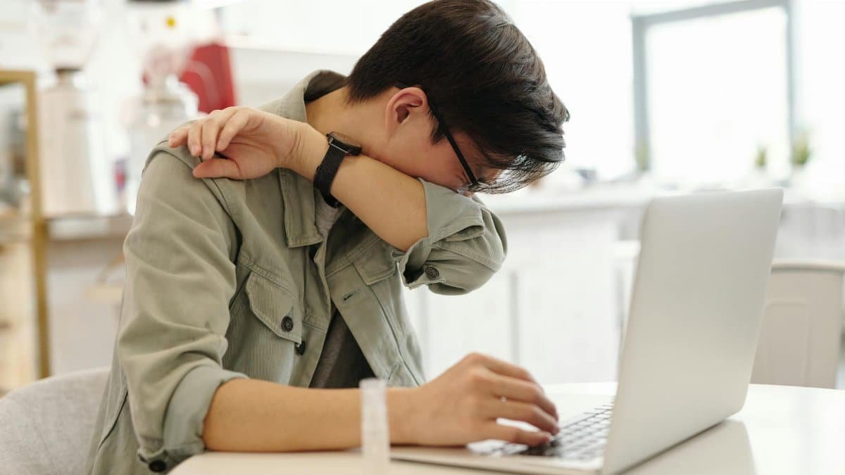 A man sneezes into his elbow while using a laptop, depicting common cold symptoms in a modern workspace.