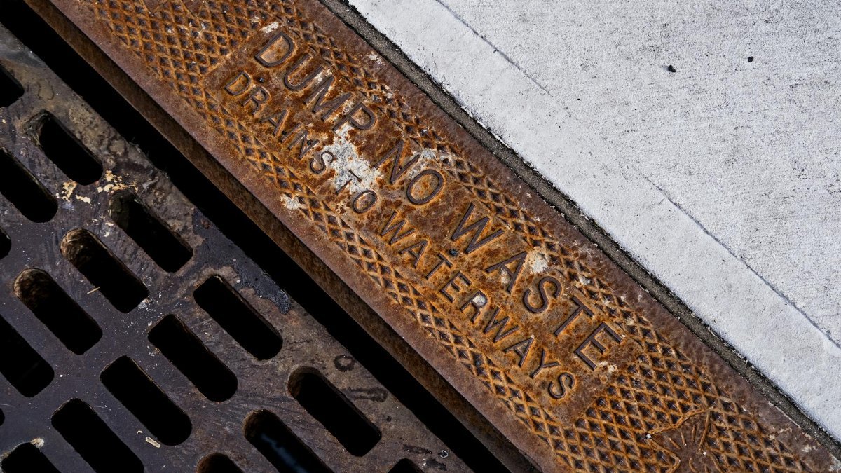 Close-up of a rusty drainage grate with environmental warning text on concrete sidewalk
