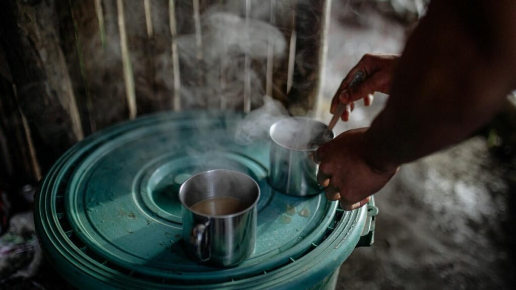 A person stirs coffee in steaming steel mugs on a green surface indoors.