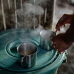 A person stirs coffee in steaming steel mugs on a green surface indoors.