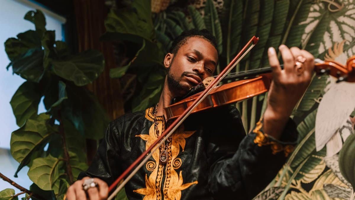 African American man playing violin, wearing traditional clothing, surrounded by indoor plants.