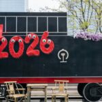 A festive train decorated with '2026' and a red bow, set in an urban environment.