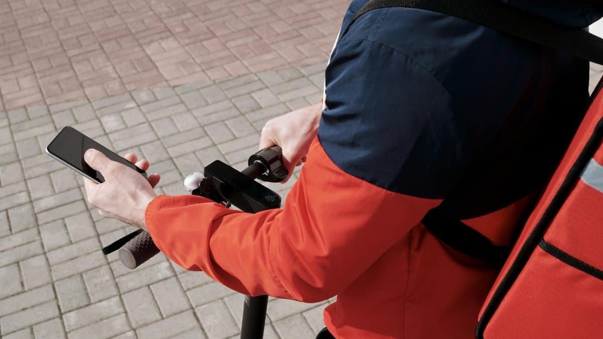 A delivery person riding an electric scooter using a smartphone for navigation outdoors.