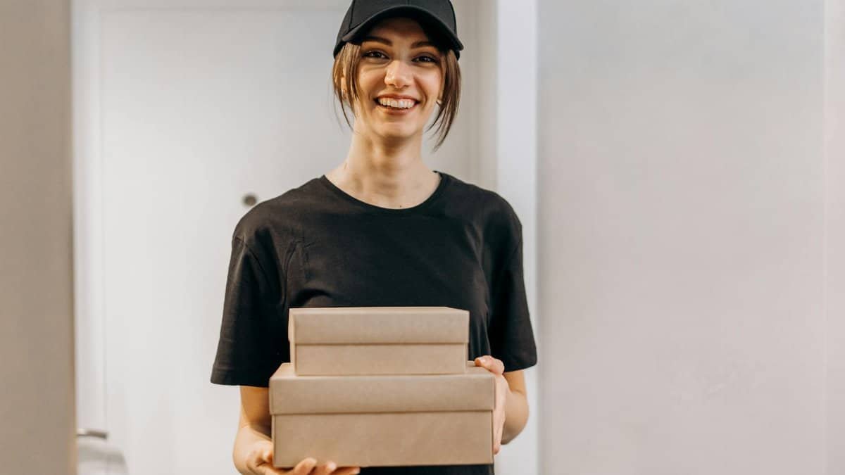 Cheerful delivery woman holding packages indoors, providing online shopping service.