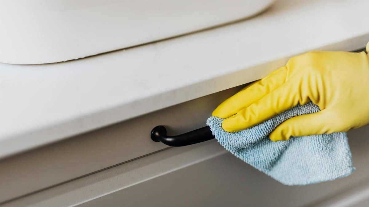 From above of crop faceless person in yellow gloves cleaning handle of cabinet with microfiber cloth in modern bright bathroom
