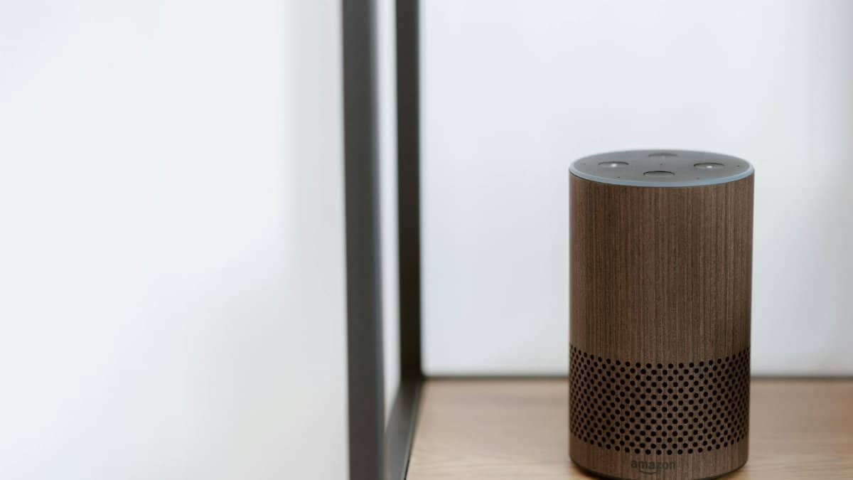 A sleek, modern smart speaker displayed on a wooden shelf in a minimalist room setting.