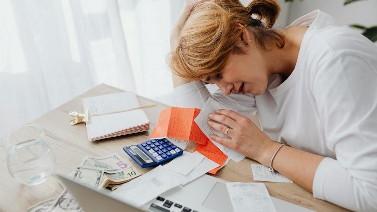 A woman overwhelmed with finances, surrounded by bills, calculator, and cash at home.