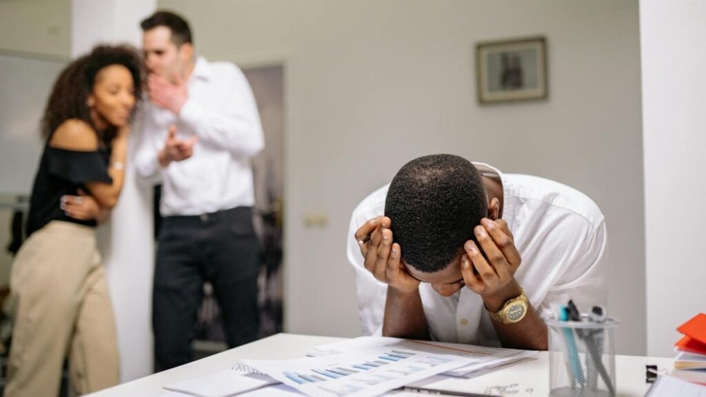 Despairing worker at office desk as colleagues gossip in background illustrating workplace bullying.