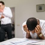 Despairing worker at office desk as colleagues gossip in background illustrating workplace bullying.
