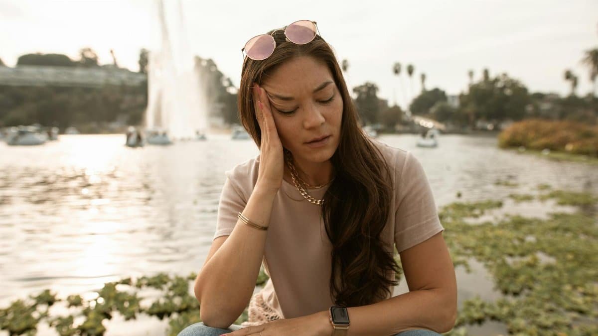 Woman feeling stressed and anxious, sitting by a lake with a serene background.