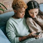 Concentrated young African American female scrolling feed in social network while resting on couch with interested Hispanic female