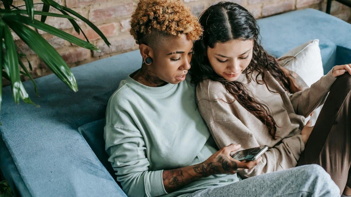 Concentrated young African American female scrolling feed in social network while resting on couch with interested Hispanic female