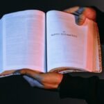 A person holds an open Bible showcasing the New Testament under dramatic lighting.