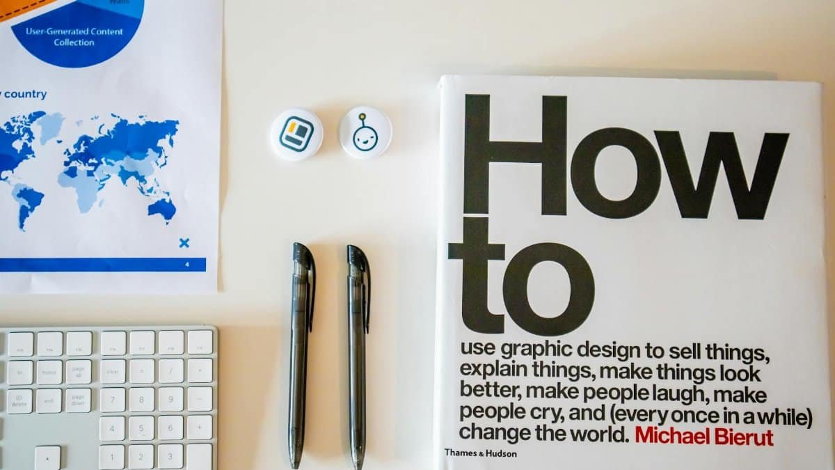 Flat lay of a creative workspace with 'How To' book, chart, pens, and keyboard in minimalist style.