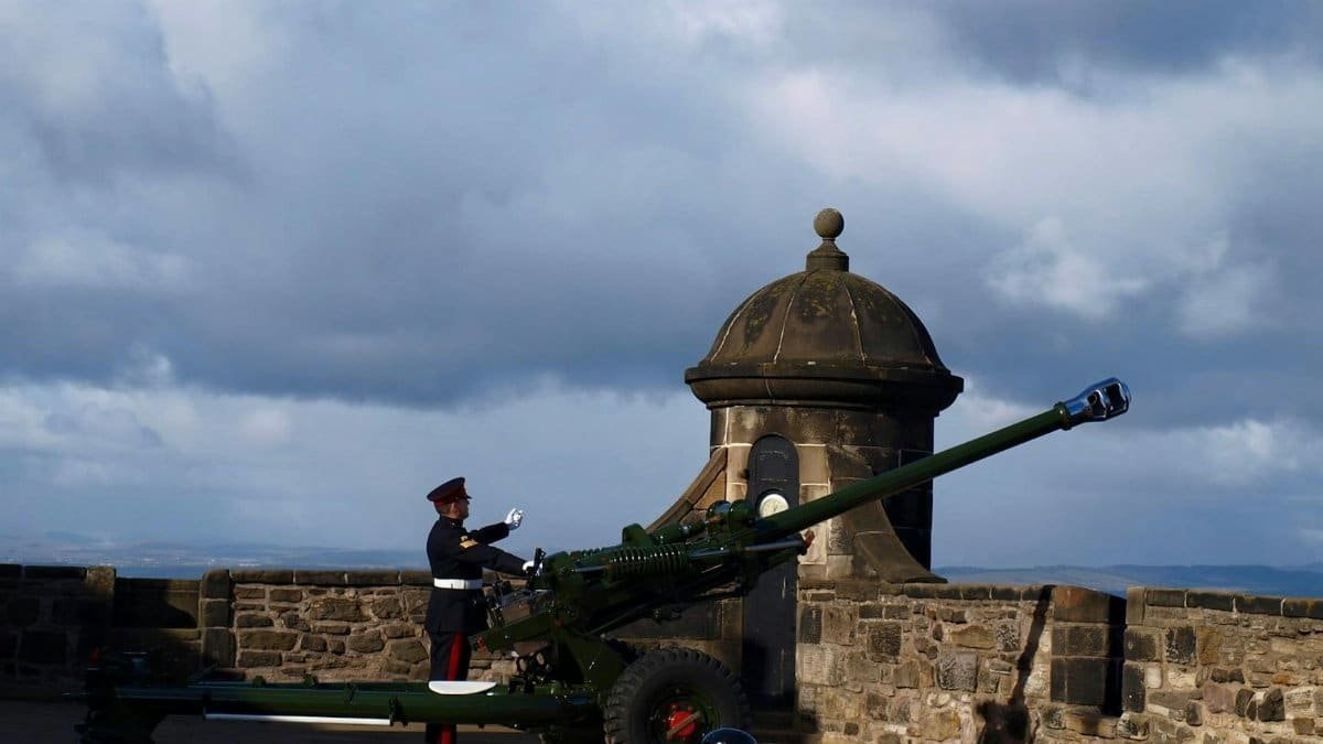 A soldier in uniform stands beside a historic cannon on a fortress wall against a cloudy sky.