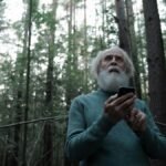 Senior man looking upwards in a forest, holding a mobile phone, surrounded by trees.