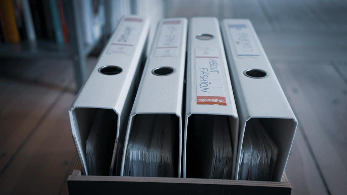 Four labeled office binders organized neatly on a wooden shelf in an indoor setting.