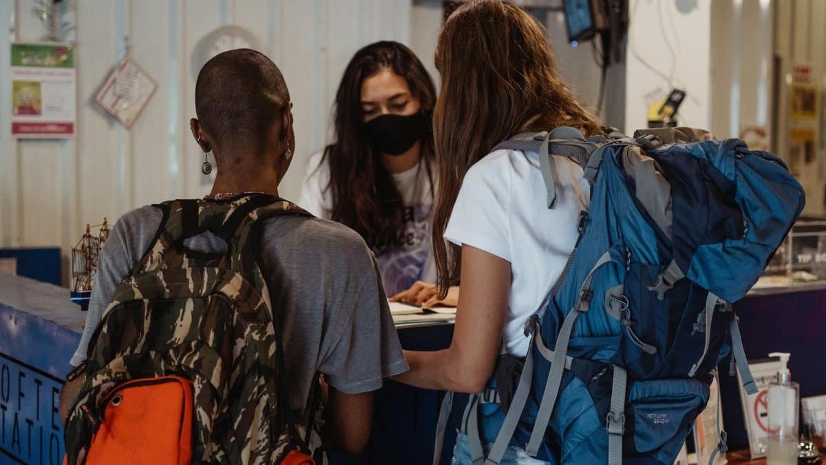Two backpackers checking in at a hostel reception, wearing face masks.