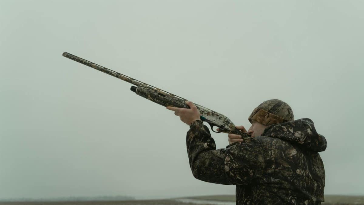 Hunter in camouflage aiming a shotgun in an open field, ready for hunting.