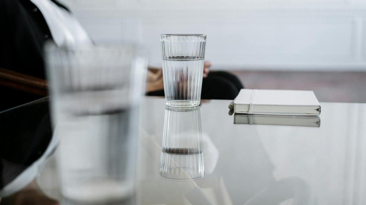 A minimalist composition featuring a glass of water and notebook on a reflective glass table.
