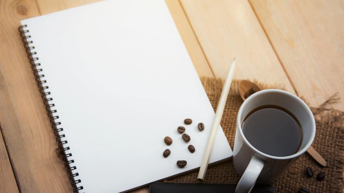 Spiral notebook, coffee cup, pencil, and smartphone on wooden desk, ideal for office or creative work concepts.
