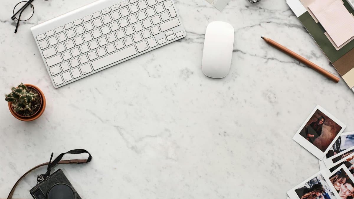 Top view of a minimalist workspace with a keyboard, camera, and instant photos on a marble desk.