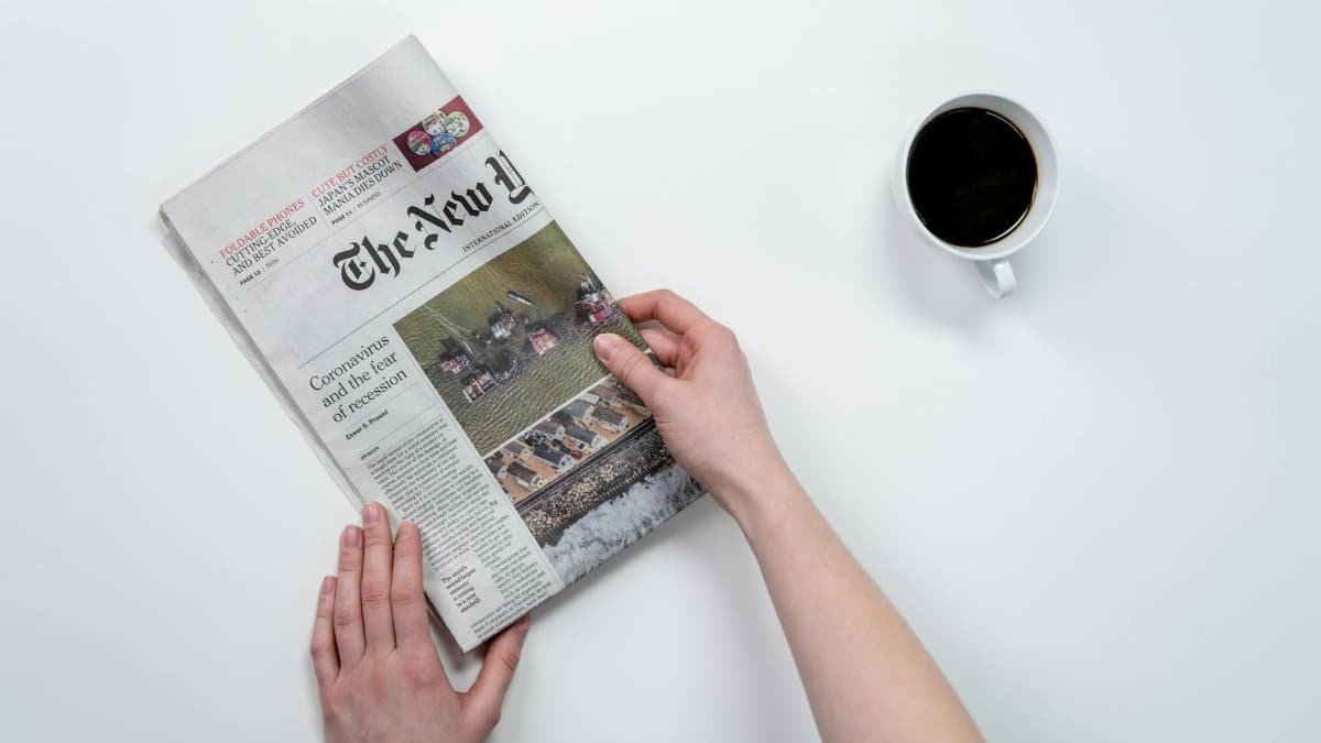 Flat lay of hands holding The New York Times newspaper with coffee on a white table.