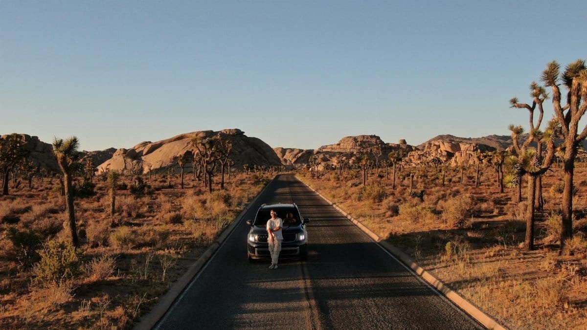 A man leans against a car on a road surrounded by Joshua trees in a desert landscape during sunset.