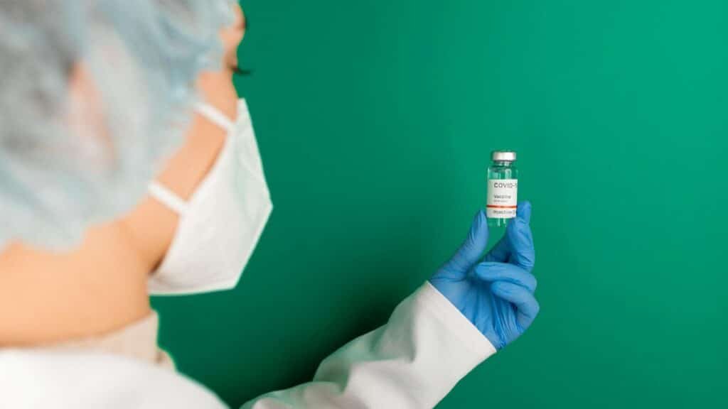 A healthcare worker wearing gloves and a mask holds a COVID-19 vaccine vial against a green background.