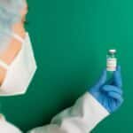 A healthcare worker wearing gloves and a mask holds a COVID-19 vaccine vial against a green background.