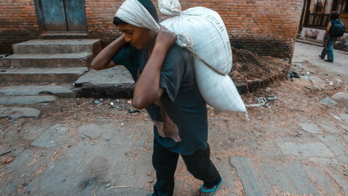 An adult man engaged in manual labor, carrying a heavy sack in an outdoor urban setting.