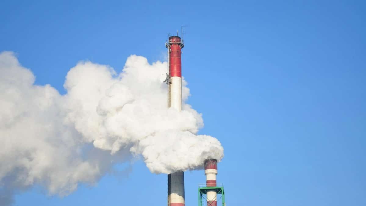A tall industrial smokestack releasing thick white smoke against a clear blue sky.