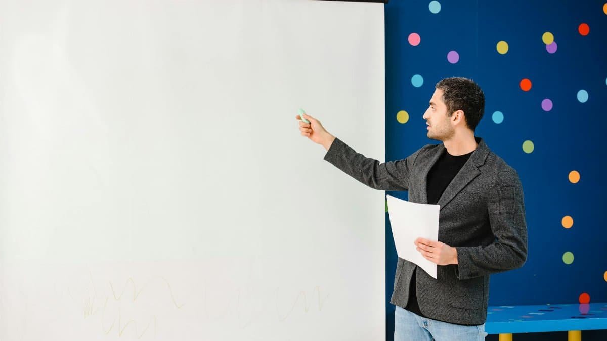 Educator in gray blazer presenting and pointing at a blank projector screen.