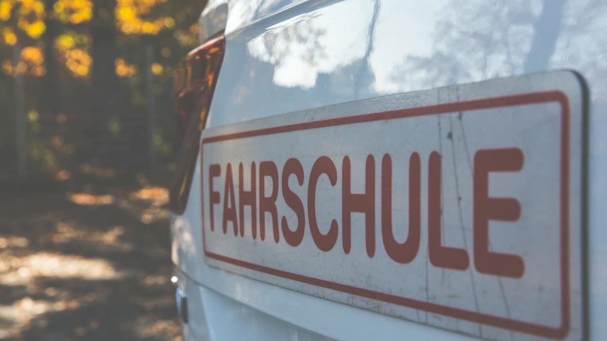 A side view of a white car with a Fahrschule sign, outdoors with autumn foliage.