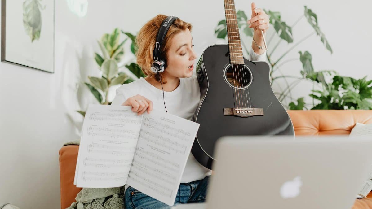A woman with headphones teaches guitar online, holding sheet music and an acoustic guitar.